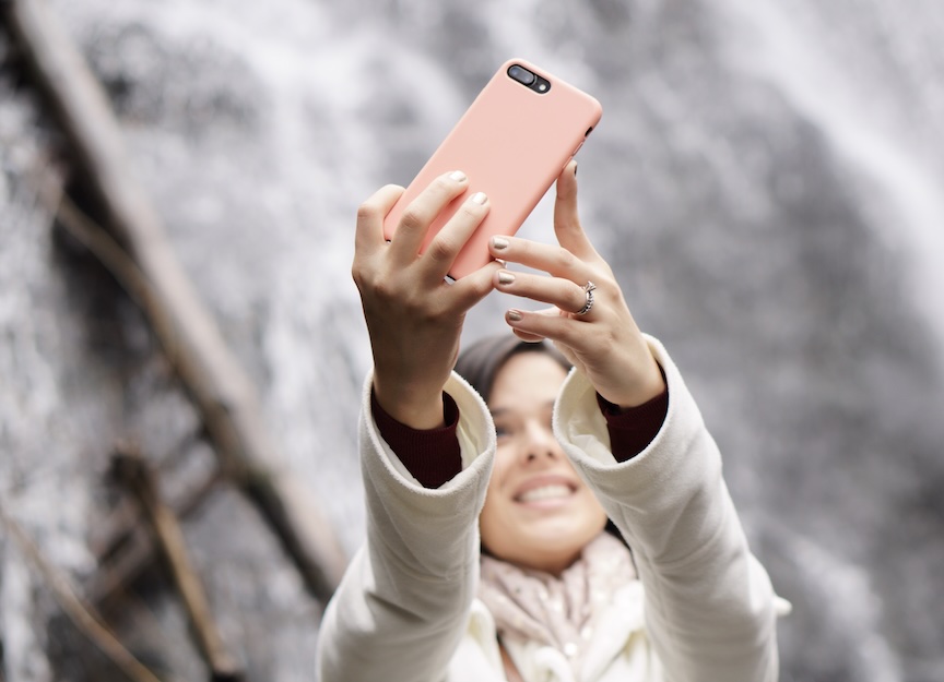 A woman holding up a smartphone to take a selfie in front of a waterfall