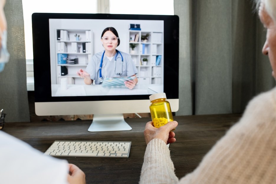 An older patient holding a pill bottle during a video visit with a doctor shown on a desktop computer screen