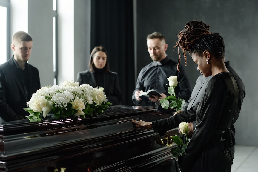 Mourners in black standing at a casket covered with white roses during a funeral service