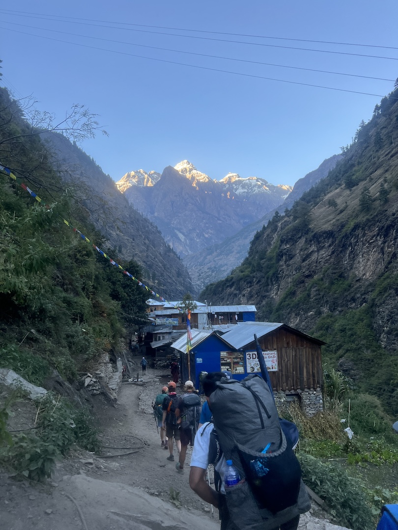 Trekkers walking toward a lodge village on the Manaslu Circuit at first light, prayer flags overhead